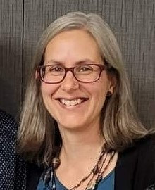  Leah has shoulder length, straight silver hair and wears purple framed glasses.